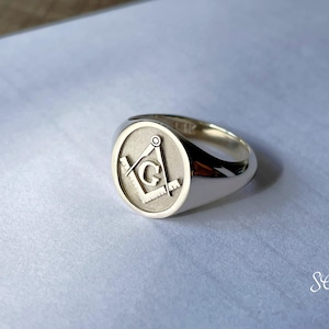May include: A silver signet ring with a Masonic symbol engraved on the top. The symbol is a square and compass with a 'G' in the center.
