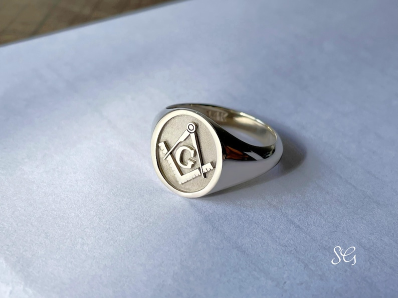 May include: A silver signet ring with a Masonic symbol engraved on the top. The symbol is a square and compass with a 'G' in the center.