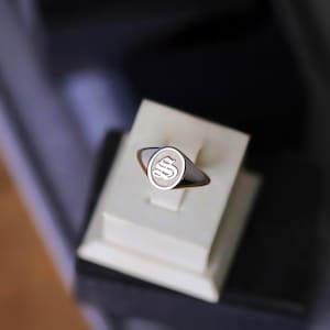 May include: A silver signet ring with a polished band and an oval face. The face features the capital letter "S" in a stylized, engraved design. The ring is displayed on a white stand.