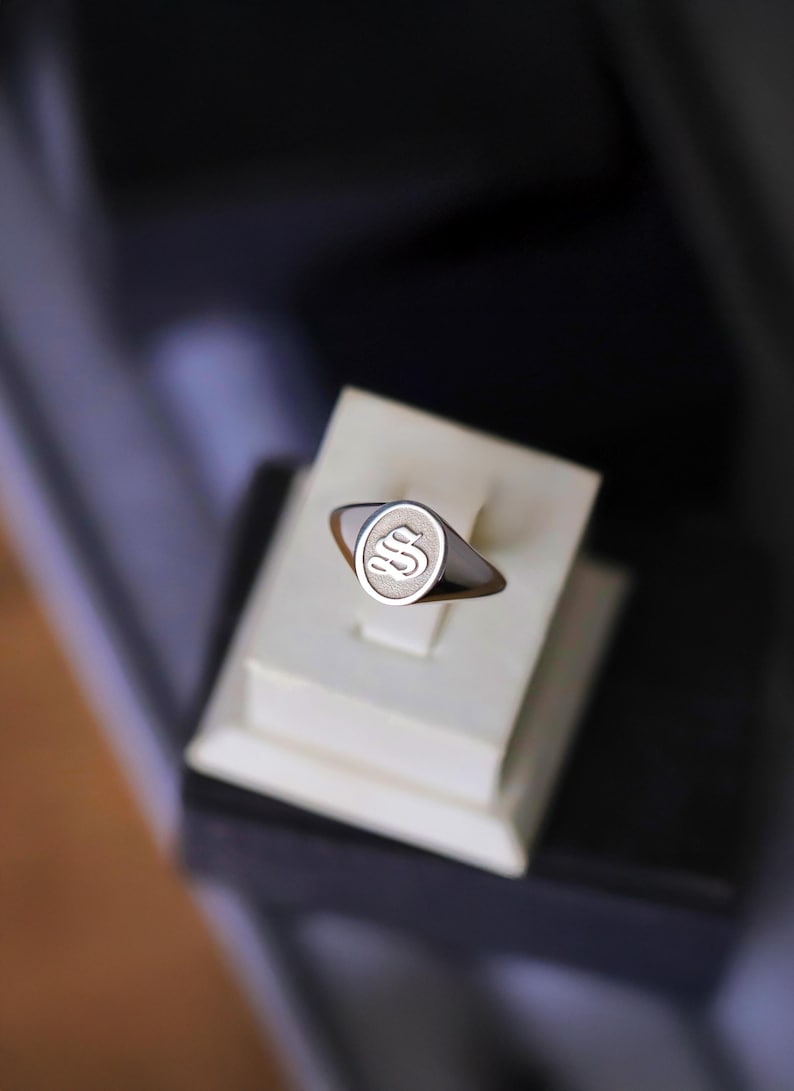 May include: A silver signet ring with a polished band and an oval face. The face features the capital letter "S" in a stylized, engraved design. The ring is displayed on a white stand.