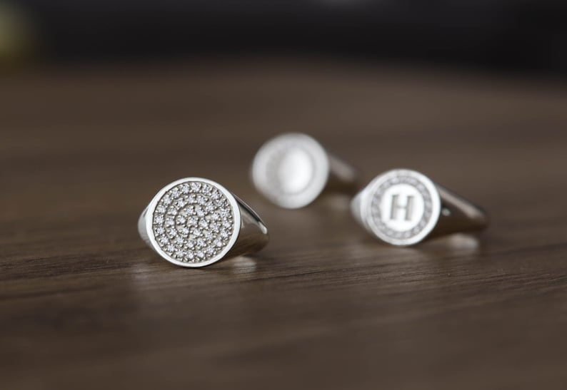 May include: Three silver signet rings with a round face. The ring on the left has a face covered in small, clear stones. The ring in the middle has a face with a single letter "O" engraved in the center. The ring on the right has a face with a single letter "H" engraved in the center.