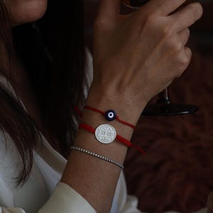 Puede incluir: Una muñeca de mujer con dos pulseras de cuerda roja. Una pulsera tiene un dije de plata con un diseño y la otra tiene un dije azul de ojo turco.