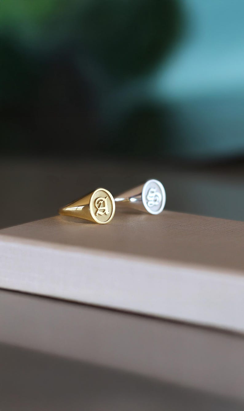 May include: Two signet rings, one gold and one silver, are shown on a neutral surface. The gold ring has the letter "A" in a serif font, while the silver ring has a stylized design. Both rings have a classic, rounded shape.