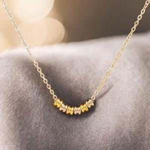 May include: Delicate gold-toned necklace featuring a series of small, circular beads. The beads alternate between gold and silver colors, creating a subtle contrast. The necklace is displayed against a soft, neutral background, highlighting its elegant design.