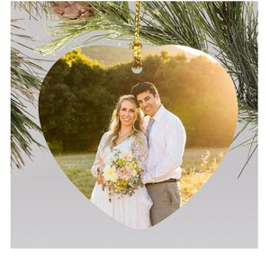 May include: Heart-shaped ornament featuring a couple embracing, set against a sunlit outdoor backdrop. The woman wears a white dress and holds a bouquet. The ornament is suspended by a gold string, with pine needles and a pinecone in the background.