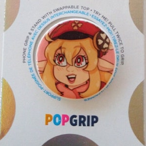 May include: A white and pink PopSockets PopGrip phone grip with a cartoon image of a girl with blonde hair wearing a red hat. The text "PopGrip" is printed in pink, blue, and green letters on the bottom of the grip.