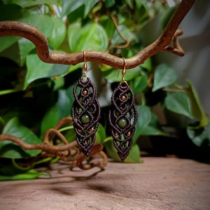 Peut inclure: Une paire de boucles d'oreilles en macramé marron foncé présentant un motif noué complexe en forme de feuille. Chaque boucle est ornée d'une pierre précieuse ronde verte au centre et de petites perles dorées, suspendues à des crochets dorés.