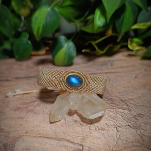 Könnte beinhalten: Ein beiges, gewebtes Makramee-Armband mit einem zentralen, ovalen Labradorit-Edelstein in einer goldfarbenen Metallfassung. Das Armband hat ein Fischgrätenmuster und verfügt über verstellbare Kordelenden mit kleinen Perlen an den Spitzen.