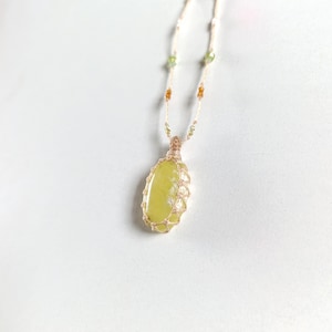 May include: A necklace featuring a smooth, oval-shaped lime green stone encased in a light-colored woven cord cage. The cord continues upward, decorated with small, alternating green and amber-colored beads along the length of the necklace chain.