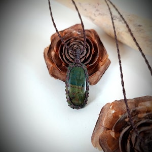 May include: A handcrafted necklace featuring a green and blue stone pendant encased in a woven brown cord. The pendant hangs from a matching cord, displayed against a backdrop of dried, brown pine cones and a white surface.