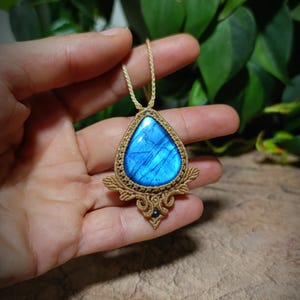 May include: A teardrop-shaped labradorite pendant featuring a vibrant blue iridescent stone set in intricate tan macrame cord work. The pendant includes a small dark bead accent at the base and hangs from a matching tan cord necklace.