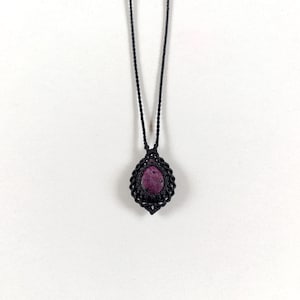 Puede incluir: Un collar con una piedra en forma de lágrima de color magenta oscuro engastada en un colgante de cordón de macramé negro. El colgante cuelga de un cordón negro trenzado fino que se extiende hacia arriba, mostrando un nudo complejo alrededor.