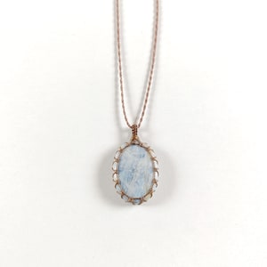 Op de afbeelding: Een handgemaakte ketting met een ovale, lichtblauwe edelsteen hanger. De hanger is gevat in een koperkleurig draadframe en hangt aan een bijpassend koord. De edelsteen heeft een gemarmerd uiterlijk, tegen een witte achtergrond.