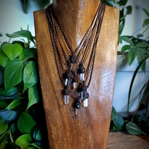 Peut inclure: Un buste en bois présente plusieurs colliers avec pendentifs en cristal. Chaque collier est orné d'un cristal de quartz clair suspendu à un cordon marron avec des nœuds décoratifs et de petites perles. Le buste est placé devant un fond de verdure.