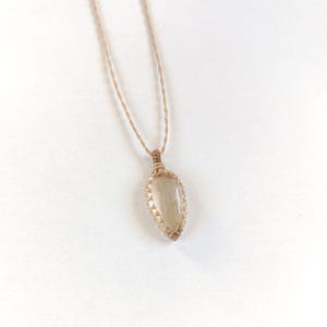 Könnte beinhalten: Eine zarte Halskette mit einem hellgrauen, tropfenförmigen Steinanhänger, der in ein geflochtenes Drahtdesign eingefasst ist. Die Halskette besteht aus einer dünnen, hellbraunen Schnur.