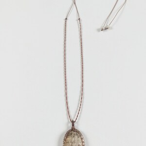 May include: A long, brown cord necklace with an oval pendant. The pendant features a light-colored stone with brown and gray inclusions, encased in a woven, metallic frame. The necklace is displayed against a white background.