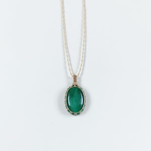 Puede incluir: Un collar con un colgante de piedra de malaquita verde con un cordón anudado. El colgante tiene forma ovalada y un color verde natural.