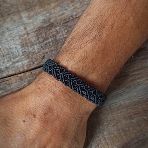 May include: A dark grey macramé bracelet with a woven geometric pattern. The bracelet is worn on a wrist, showcasing the intricate knotting detail. The bracelet is made of a dark grey cord.