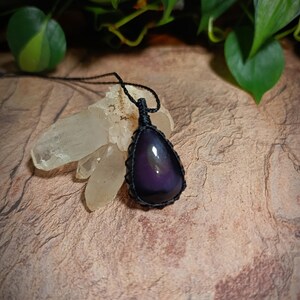 May include: A teardrop-shaped purple and black stone pendant secured in a black macrame cord setting. The pendant hangs from a thin black cord and is positioned against a cluster of clear, jagged quartz crystals with a smooth, polished surface.