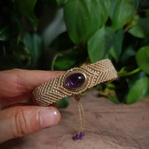 May include: A beige macrame bracelet featuring a central oval-shaped purple amethyst gemstone set in a gold-toned metal bezel. The woven cord band displays a chevron pattern and includes two dangling strings finished with small purple beads.