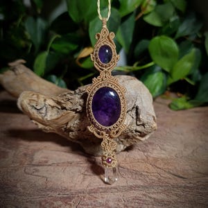 May include: A macrame pendant necklace featuring a tan cord woven into intricate patterns around two oval purple amethyst stones and a small clear quartz crystal point at the base, accented with a tiny red bead above the crystal point.