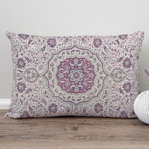 May include: A patterned cushion in purple and cream tones, featuring a central floral design. It rests on a wooden surface, accompanied by a decorative ball and a small pot of lavender.
