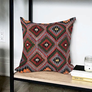 Op de afbeelding: Vierkant decoratief kussen met een geometrisch patroon in rood, groen, grijs en oranje. Het kussen heeft een ruitvormig ontwerp met ingewikkelde details, wat kleur en textuur toevoegt.