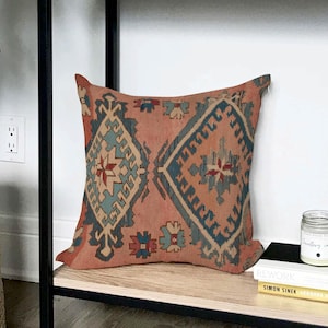 Puede incluir: Un cojín decorativo cuadrado con un patrón geométrico en tonos coral, turquesa y beige. El cojín está colocado sobre una estantería de madera, añadiendo un toque bohemio a la habitación. El diseño presenta motivos de diamantes y flores.