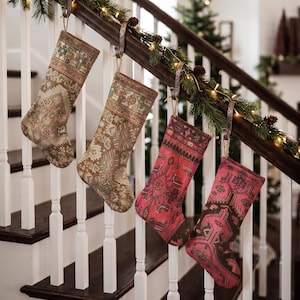 May include: Four Christmas stockings hanging from a wooden staircase railing. Two stockings feature muted earth tones with intricate geometric patterns, while the other two display vibrant pink and dark brown hues with similar detailed motifs.