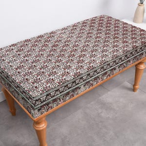 May include: A wooden bench with a patterned cushion. The cushion is cream coloured with a black and red floral design. The bench has four legs and is made of wood.