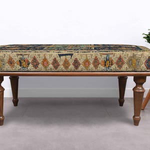 May include: A rectangular ottoman with a patterned top and dark brown wooden legs. The top features a beige background with a repeating pattern of colourful geometric shapes. The legs are turned and taper towards the floor, adding a touch of elegance.