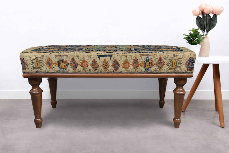 May include: A rectangular ottoman with a patterned top and dark brown wooden legs. The top features a beige background with a repeating pattern of colourful geometric shapes. The legs are turned and taper towards the floor, adding a touch of elegance.