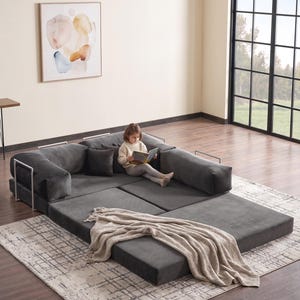 May include: A modular, dark gray sectional sofa with white metal arm supports. A child is sitting on the sofa, reading a book. A beige throw blanket is draped over the sofa. The sofa sits on a patterned rug.
