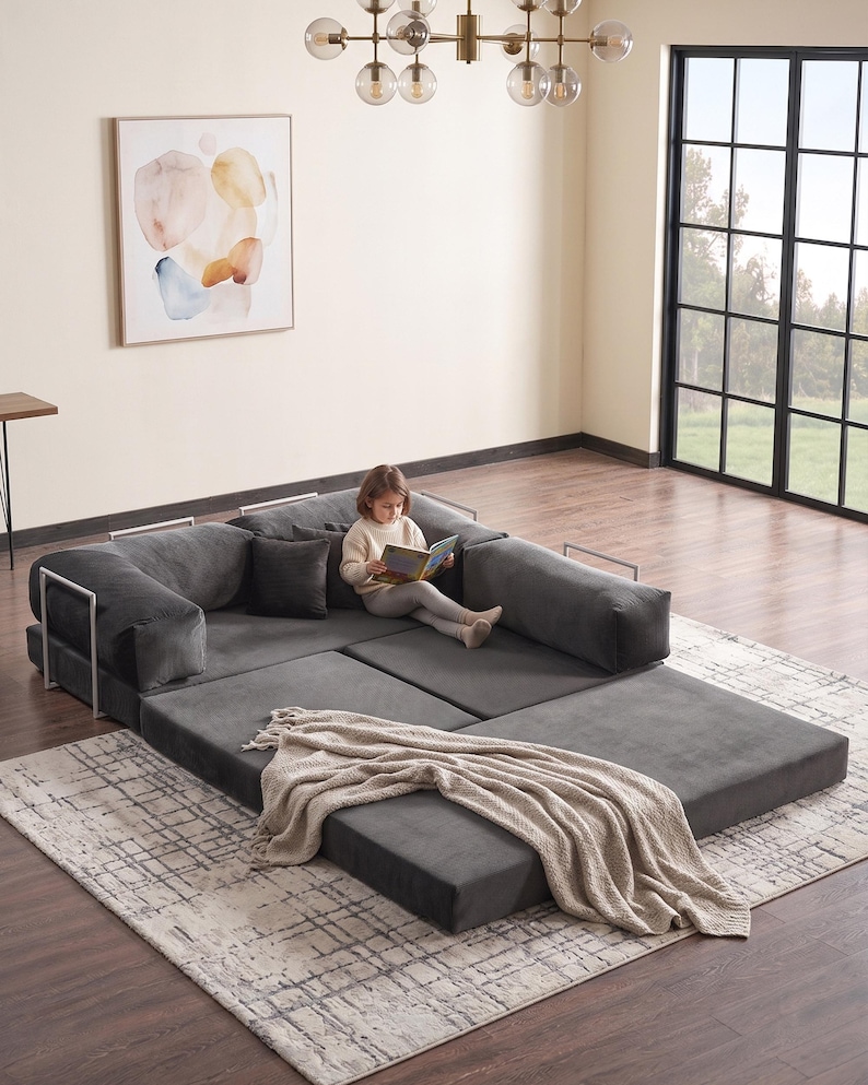 May include: A modular, dark gray sectional sofa with white metal arm supports. A child is sitting on the sofa, reading a book. A beige throw blanket is draped over the sofa. The sofa sits on a patterned rug.