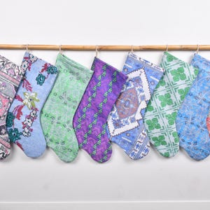 Puede incluir: Una colección de ocho calcetines navideños con varios patrones y colores, incluyendo diseños florales, geométricos y paisley. Los calcetines están suspendidos de una vara de bambú, creando una exhibición festiva. La paleta de colores incluye rosa, azul, verde y morado.