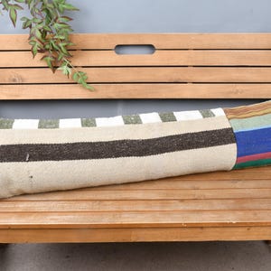 May include: A long, rectangular decorative pillow with a patchwork design. The pillow features a beige base with brown stripes, green and white blocks, and a colorful striped end. It rests on a wooden bench.