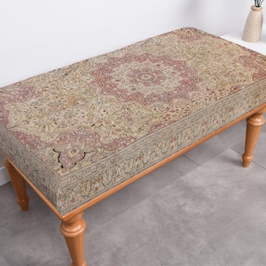 May include: A wooden bench with a patterned fabric cushion. The cushion features a floral design in shades of beige, brown, and pink. The bench has four legs and is made of wood.