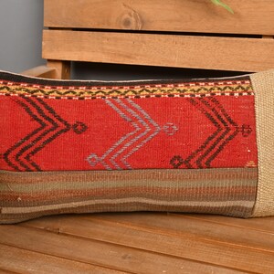 May include: Rectangular decorative pillow with a vibrant red central panel featuring geometric patterns in black and gray. The pillow has beige end panels and a striped lower section, resting on a wooden surface.