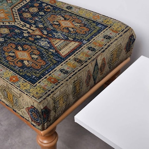 May include: A rectangular ottoman with a patterned fabric top. The fabric features a traditional rug design with blue, orange, and beige colours. The ottoman has wooden legs and a sturdy frame, suitable for seating or as a decorative piece.