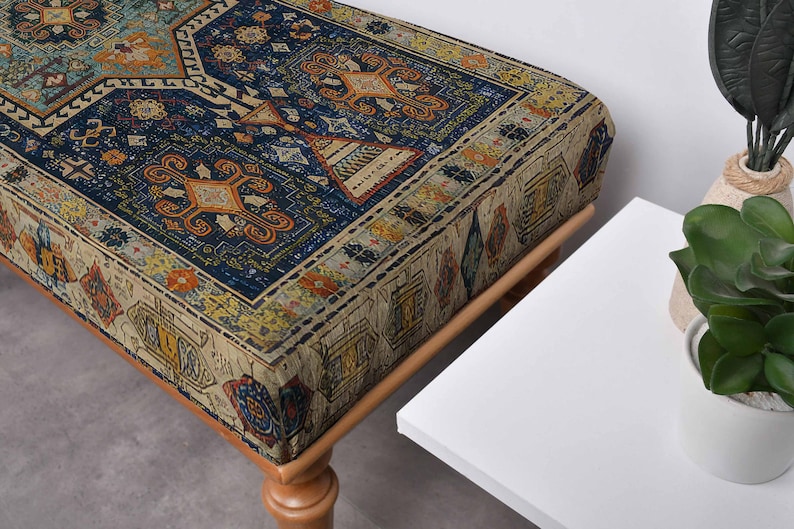May include: A rectangular ottoman with a patterned fabric top. The fabric features a traditional rug design with blue, orange, and beige colours. The ottoman has wooden legs and a sturdy frame, suitable for seating or as a decorative piece.