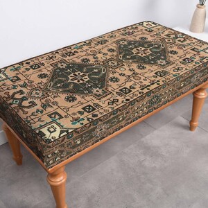 May include: A rectangular bench with a patterned top and wooden legs. The bench's top features a beige and dark green design with geometric shapes. The legs are light brown. The bench is a decorative furniture piece.