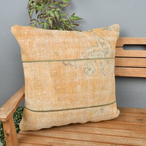 May include: A light brown throw pillow with a subtle pattern and a green border. The pillow is sitting on a wooden bench.