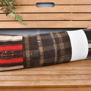 May include: A long, rectangular cushion with a patchwork design. The cushion features sections of red and beige stripes, brown plaid, a white stripe, and dark stripes. It is placed on a wooden surface.