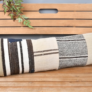 May include: Rectangular decorative pillow with a patchwork design. The pillow features sections of beige, white, and brown fabric with striped and solid patterns. The pillow is placed on a wooden bench.