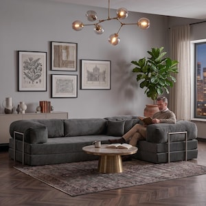 Puede incluir: Un sal&oacute;n moderno con un gran sof&aacute; seccional gris oscuro con detalles met&aacute;licos. Una mesa de centro redonda con tapa de m&aacute;rmol y base dorada descansa sobre una alfombra estampada. Obras de arte cuelgan de la pared gris y una l&aacute;mpara decorativa cuelga del techo.