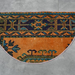 Puede incluir: Una alfombra de media luna con un patrón geométrico en tonos naranja, azul, verde y negro. La alfombra tiene un fleco rosa alrededor de los bordes.