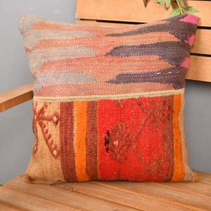 May include: A square, decorative pillow with a woven, textured design. The top portion features horizontal stripes in shades of peach, gray, and purple. The bottom section is primarily red with orange and tan vertical stripes and geometric patterns.