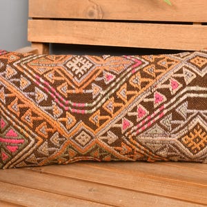 May include: Rectangular decorative pillow with a brown background and a geometric pattern in shades of orange, pink, and beige. The pillow is placed on a wooden bench, showcasing its intricate design and texture. A stylish home decor item.
