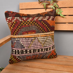 May include: A decorative square pillow with a woven, patterned design in shades of red, orange, brown, and purple. The pillow is placed on a wooden bench, showcasing its texture and color variations. The pillow's dimensions are approximately 16 inches by 16 inches.