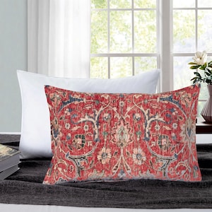 Pode incluir: Uma almofada retangular decorativa com um padrão floral vermelho, azul e creme. A almofada tem um aspeto vintage e desgastado e é colocada numa superfície cinzenta escura. Uma almofada branca está atrás.